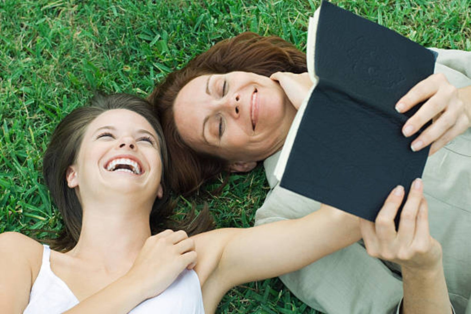 Two women lying in the grass are laughing with each other while reading a book.