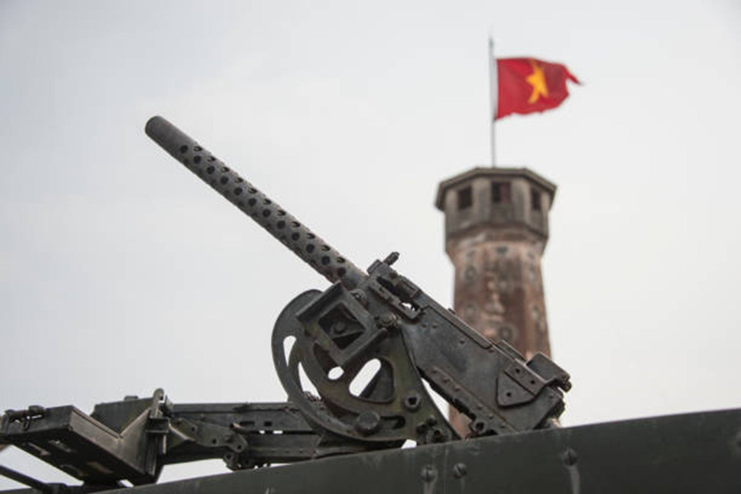 Close-up of an old military machine gun with a tower and a Vietnamese flag in the background.